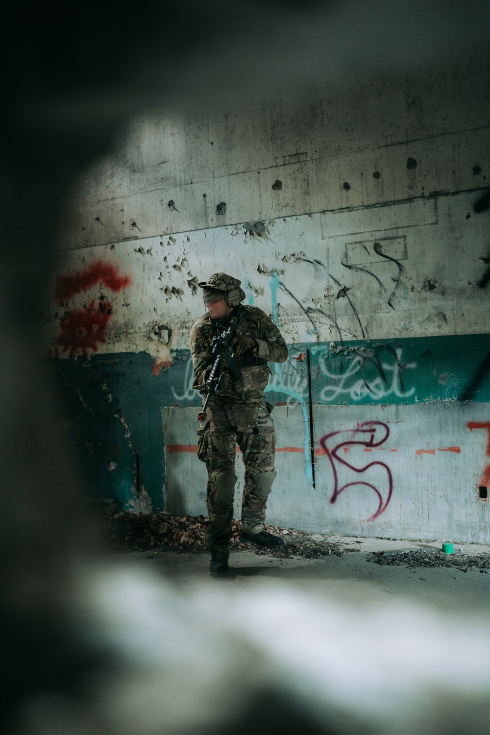 A heavily equipped individual in full camouflage tactical gear stands inside a deteriorated building with heavily damaged walls. The person is holding a rifle and wearing tactical headgear, photographed through a gap or hole that frames the scene. The surrounding walls display extensive graffiti, pockmarks, and peeling paint suggesting an abandoned or derelict urban structure.