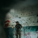 A heavily equipped individual in full camouflage tactical gear stands inside a deteriorated building with heavily damaged walls. The person is holding a rifle and wearing tactical headgear, photographed through a gap or hole that frames the scene. The surrounding walls display extensive graffiti, pockmarks, and peeling paint suggesting an abandoned or derelict urban structure.