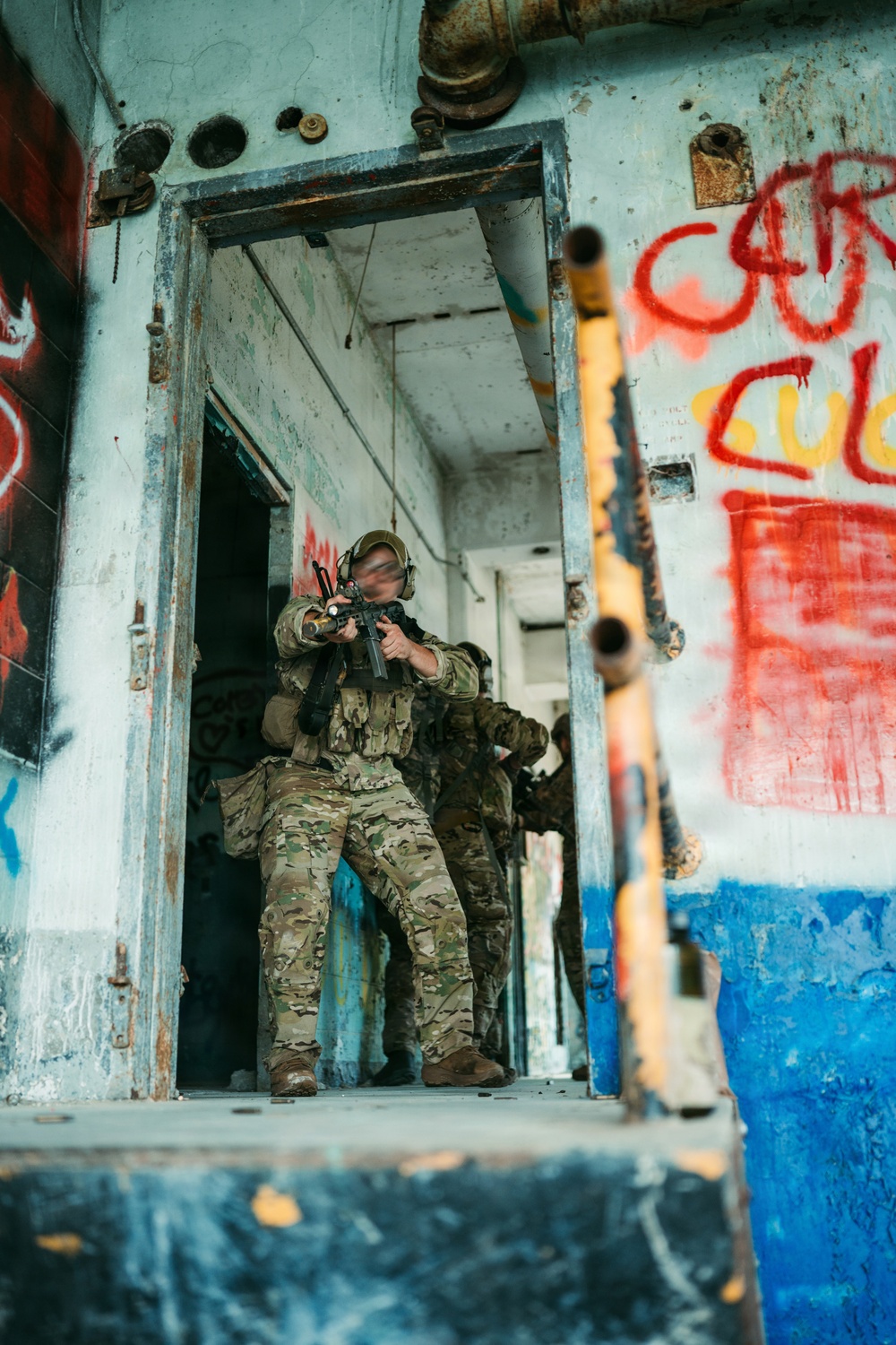 The image shows one or more individuals in full military/tactical camouflage gear (MultiCam pattern) conducting what appears to be a building-clearing or close-quarters battle (CQB) drill inside a dilapidated, graffiti-covered structure. The lead figure is positioned in a doorway with a rifle raised in a firing stance. The setting resembles an urban training facility or abandoned industrial building.
