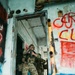 The image shows one or more individuals in full military/tactical camouflage gear (MultiCam pattern) conducting what appears to be a building-clearing or close-quarters battle (CQB) drill inside a dilapidated, graffiti-covered structure. The lead figure is positioned in a doorway with a rifle raised in a firing stance. The setting resembles an urban training facility or abandoned industrial building.