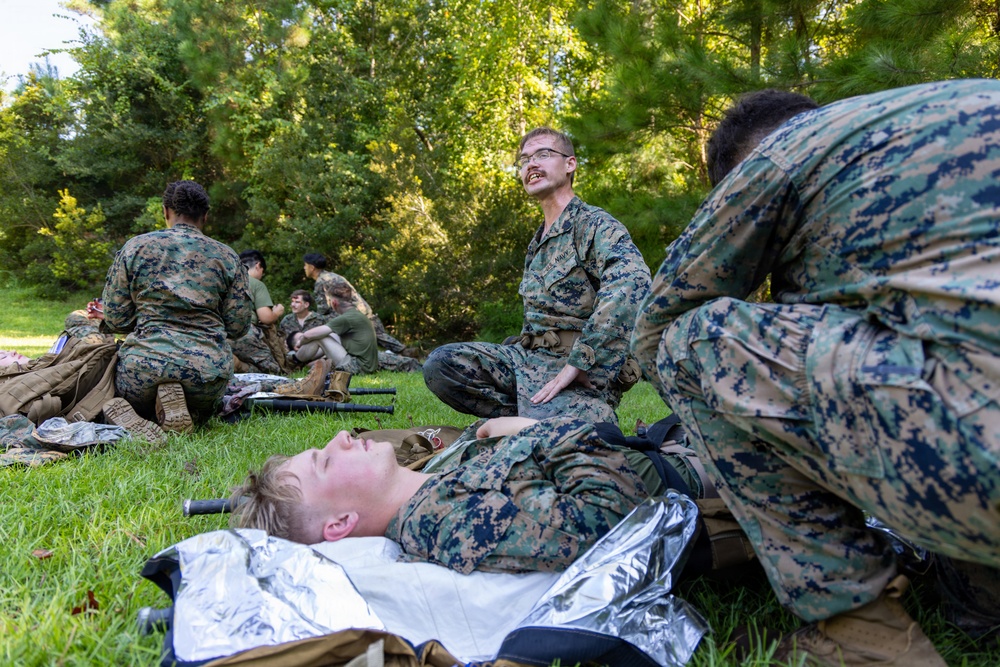 Military personnel in MARPAT camouflage uniforms are conducting what appears to be a combat casualty care or field medical training exercise outdoors. One service member is lying on a mylar emergency blanket on the grass while others attend to him, with additional pairs of personnel practicing similar procedures in the background. The setting is a grassy area bordered by trees, consistent with a training field environment.