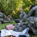 Military personnel in MARPAT camouflage uniforms are conducting what appears to be a combat casualty care or field medical training exercise outdoors. One service member is lying on a mylar emergency blanket on the grass while others attend to him, with additional pairs of personnel practicing similar procedures in the background. The setting is a grassy area bordered by trees, consistent with a training field environment.