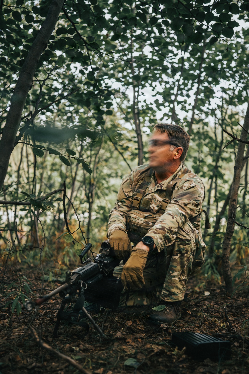 A soldier in full multicam camouflage uniform is seated in a wooded area, holding a rifle with an optic sight mounted on a bipod. The individual's face has been deliberately blurred, suggesting operational security considerations. The setting appears to be a forest or treeline during daylight hours.