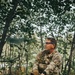 A soldier in full multicam camouflage uniform is seated in a wooded area, holding a rifle with an optic sight mounted on a bipod. The individual's face has been deliberately blurred, suggesting operational security considerations. The setting appears to be a forest or treeline during daylight hours.