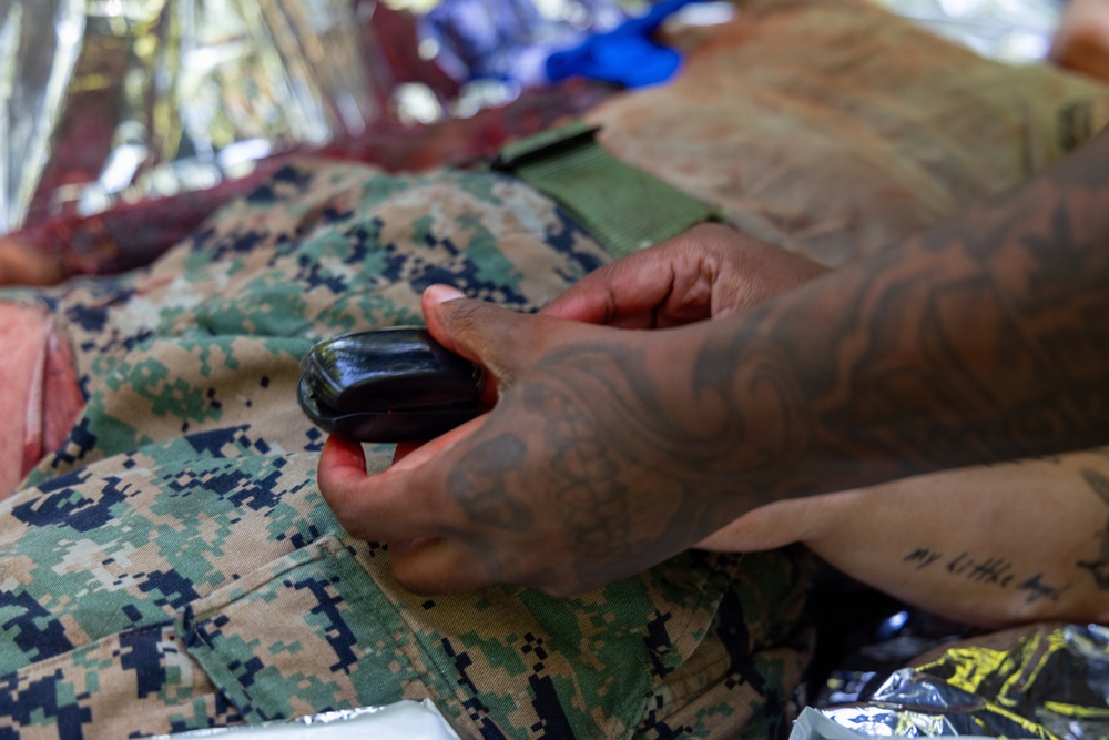 A tattooed individual is handling a small dark object over what appears to be USMC MARPAT digital camouflage uniform or gear. The scene includes what appears to be medical or tactical equipment in the background, including a mylar/emergency blanket. The context suggests a tactical combat casualty care (TCCC) or field medical training scenario.