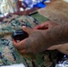 A tattooed individual is handling a small dark object over what appears to be USMC MARPAT digital camouflage uniform or gear. The scene includes what appears to be medical or tactical equipment in the background, including a mylar/emergency blanket. The context suggests a tactical combat casualty care (TCCC) or field medical training scenario.