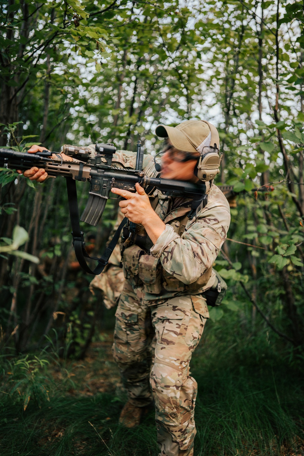 A person in full multicam camouflage tactical gear is aiming an assault rifle (appears to be an AR-15/M4-style platform) while moving through a dense forest environment. A second similarly equipped individual is visible in the background. The setting and equipment suggest a military training exercise, airsoft/milsim event, or tactical training scenario.