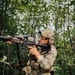 A person in full multicam camouflage tactical gear is aiming an assault rifle (appears to be an AR-15/M4-style platform) while moving through a dense forest environment. A second similarly equipped individual is visible in the background. The setting and equipment suggest a military training exercise, airsoft/milsim event, or tactical training scenario.