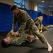 Military personnel in camouflage uniforms are practicing combatives/hand-to-hand combat training on a black mat floor. In the foreground, one soldier in a dominant position is controlling a grounded opponent, while additional pairs of trainees are visible practicing in the background. The setting appears to be an indoor training facility with blue wall padding.