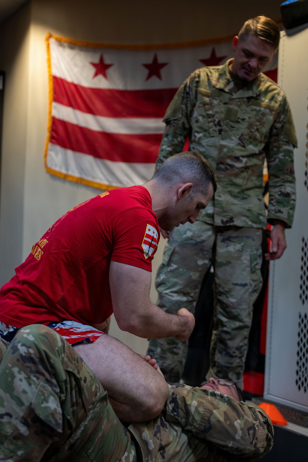 The image shows what appears to be a military combatives or ground fighting training session. One person in a red shirt and camouflage pants is in a dominant ground position over another person in full camouflage uniform, while a third person in camouflage stands and observes. The Washington D.C. flag is prominently displayed on the wall in the background.