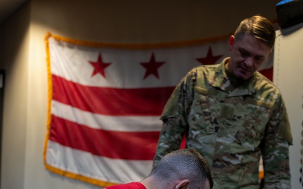 The image shows what appears to be a military combatives or ground fighting training session. One person in a red shirt and camouflage pants is in a dominant ground position over another person in full camouflage uniform, while a third person in camouflage stands and observes. The Washington D.C. flag is prominently displayed on the wall in the background.