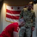 The image shows what appears to be a military combatives or ground fighting training session. One person in a red shirt and camouflage pants is in a dominant ground position over another person in full camouflage uniform, while a third person in camouflage stands and observes. The Washington D.C. flag is prominently displayed on the wall in the background.