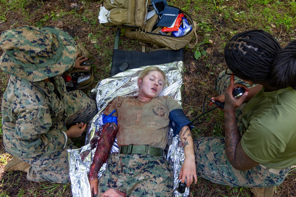 A military medical training exercise is depicted, showing a simulated casualty scenario. A female service member in Marine Corps MARPAT uniform is lying on a mylar emergency blanket with simulated wound makeup on her arm, while two other personnel provide mock medical treatment. The scene includes medical supplies and equipment consistent with a field medicine training drill.