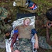 A military medical training exercise is depicted, showing a simulated casualty scenario. A female service member in Marine Corps MARPAT uniform is lying on a mylar emergency blanket with simulated wound makeup on her arm, while two other personnel provide mock medical treatment. The scene includes medical supplies and equipment consistent with a field medicine training drill.