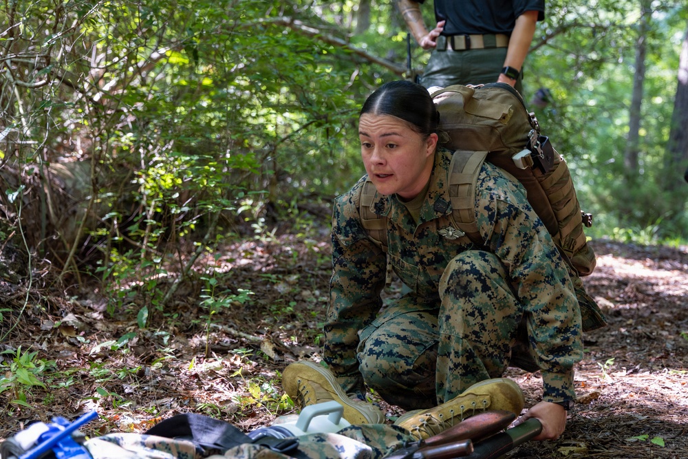A female service member in U.S. Marine Corps MARPAT woodland camouflage uniform is crouching in a forested outdoor environment, appearing to be engaged in a field training exercise. She is wearing a tactical backpack and has equipment laid out on the ground in front of her. A second individual is partially visible standing behind her.