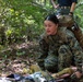 A female service member in U.S. Marine Corps MARPAT woodland camouflage uniform is crouching in a forested outdoor environment, appearing to be engaged in a field training exercise. She is wearing a tactical backpack and has equipment laid out on the ground in front of her. A second individual is partially visible standing behind her.