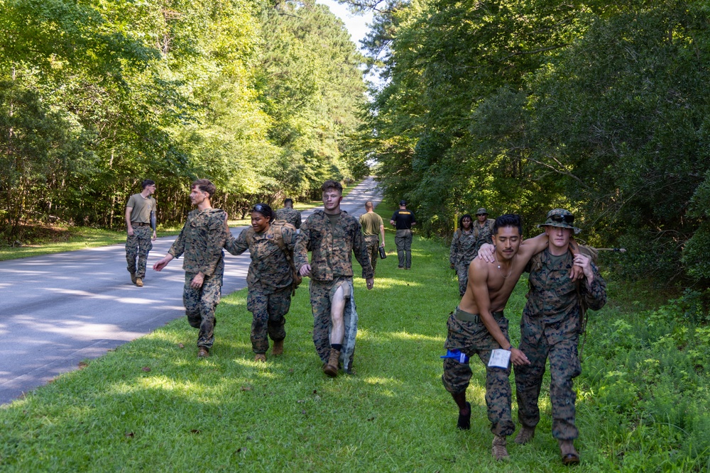 A group of individuals in U.S. Marine Corps MARPAT camouflage uniforms are conducting what appears to be a casualty evacuation or buddy-carry training exercise along a tree-lined road. In the foreground, a shirtless individual is being supported and carried by a uniformed Marine, while in the background another group of Marines appears to be assisting an individual with simulated leg injuries. The setting is a wooded area with a paved road, suggesting a military training installation.