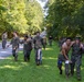 A group of individuals in U.S. Marine Corps MARPAT camouflage uniforms are conducting what appears to be a casualty evacuation or buddy-carry training exercise along a tree-lined road. In the foreground, a shirtless individual is being supported and carried by a uniformed Marine, while in the background another group of Marines appears to be assisting an individual with simulated leg injuries. The setting is a wooded area with a paved road, suggesting a military training installation.