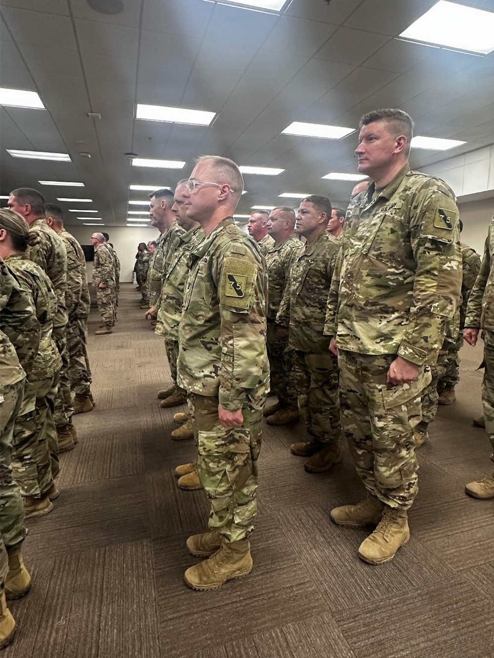 A large group of U.S. Army soldiers in OCP (Operational Camouflage Pattern) uniforms are standing in formation inside what appears to be a large indoor facility. Multiple rows of soldiers are visible, standing at ease in an organized military formation. At least two soldiers prominently display a shoulder patch with the number '75', suggesting affiliation with the 75th Division or a related unit.
