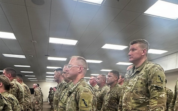 A large group of U.S. Army soldiers in OCP (Operational Camouflage Pattern) uniforms are standing in formation inside what appears to be a large indoor facility. Multiple rows of soldiers are visible, standing at ease in an organized military formation. At least two soldiers prominently display a shoulder patch with the number '75', suggesting affiliation with the 75th Division or a related unit.