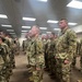 A large group of U.S. Army soldiers in OCP (Operational Camouflage Pattern) uniforms are standing in formation inside what appears to be a large indoor facility. Multiple rows of soldiers are visible, standing at ease in an organized military formation. At least two soldiers prominently display a shoulder patch with the number '75', suggesting affiliation with the 75th Division or a related unit.