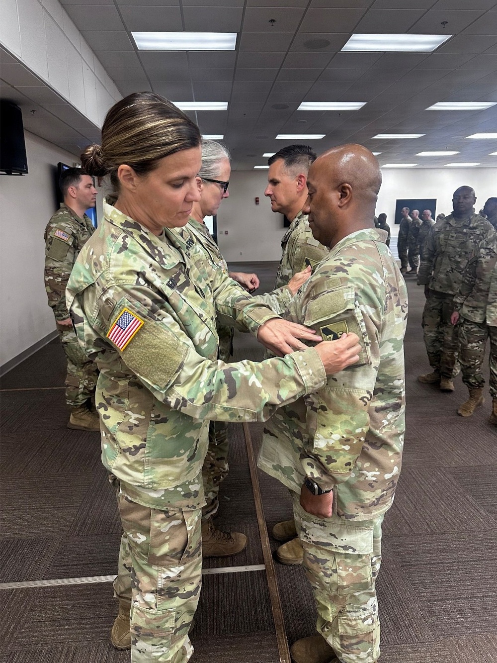 A military ceremony is taking place indoors where a female soldier is pinning something onto the chest of a male soldier, consistent with a promotion or award ceremony. Multiple soldiers in U.S. Army OCP (Operational Camouflage Pattern) uniforms are present in the background. The setting appears to be a modern military facility or conference room.
