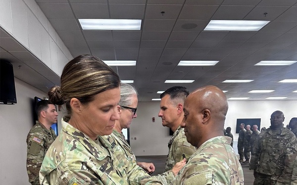 A military ceremony is taking place indoors where a female soldier is pinning something onto the chest of a male soldier, consistent with a promotion or award ceremony. Multiple soldiers in U.S. Army OCP (Operational Camouflage Pattern) uniforms are present in the background. The setting appears to be a modern military facility or conference room.