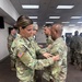 A military ceremony is taking place indoors where a female soldier is pinning something onto the chest of a male soldier, consistent with a promotion or award ceremony. Multiple soldiers in U.S. Army OCP (Operational Camouflage Pattern) uniforms are present in the background. The setting appears to be a modern military facility or conference room.