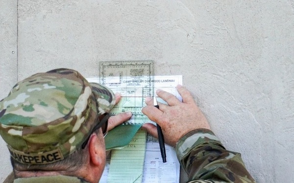 A U.S. Army soldier in OCP (Operational Camouflage Pattern) uniform is writing on or reviewing documents pinned against a concrete wall. The documents appear to be related to a 'Camp Shelby Dogwood LANDNAV' (Land Navigation) exercise. The soldier's name tape reads 'MAKEPEACE' and a unit patch is visible on the left shoulder.
