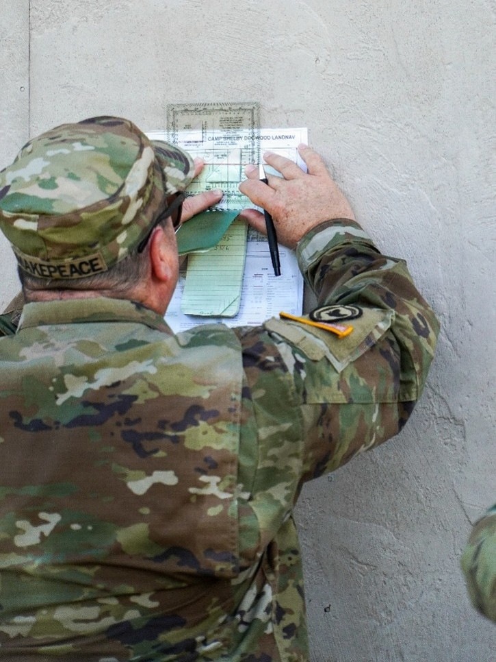 A U.S. Army soldier in OCP (Operational Camouflage Pattern) uniform is writing on or reviewing documents pinned against a concrete wall. The documents appear to be related to a 'Camp Shelby Dogwood LANDNAV' (Land Navigation) exercise. The soldier's name tape reads 'MAKEPEACE' and a unit patch is visible on the left shoulder.