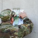 A U.S. Army soldier in OCP (Operational Camouflage Pattern) uniform is writing on or reviewing documents pinned against a concrete wall. The documents appear to be related to a 'Camp Shelby Dogwood LANDNAV' (Land Navigation) exercise. The soldier's name tape reads 'MAKEPEACE' and a unit patch is visible on the left shoulder.
