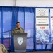 A U.S. Navy officer in uniform is speaking at a podium bearing the Surface Navy Association logo at the 2026 Waterfront Symposium West Coast event. The setting is a tent or temporary venue decorated with blue draping, with sponsor banners visible in the background. The event features prominent defense industry sponsors including General Dynamics Mission Systems, Saronic, BAE Systems, HII, and Lockheed Martin.