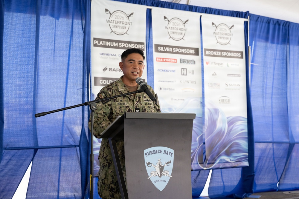 A U.S. Navy officer in digital camouflage uniform is speaking at a podium bearing the Surface Navy Association logo. The event is the 2026 Waterfront Symposium, West Coast edition, with sponsor banners visible in the background including Platinum, Silver, and Bronze sponsor categories. The setting appears to be a tent or temporary venue decorated with blue draping.