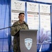 A U.S. Navy officer in digital camouflage uniform is speaking at a podium bearing the Surface Navy Association logo. The event is the 2026 Waterfront Symposium, West Coast edition, with sponsor banners visible in the background including Platinum, Silver, and Bronze sponsor categories. The setting appears to be a tent or temporary venue decorated with blue draping.