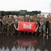 A large group of military personnel in camouflage uniforms pose for a group photograph on a wet tarmac or base facility. Two individuals in the front center are holding a red flag bearing the U.S. Army Corps of Engineers castle insignia. A building in the background is partially visible with signage reading 'Marine Wing Supp...' suggesting a Marine Corps installation.