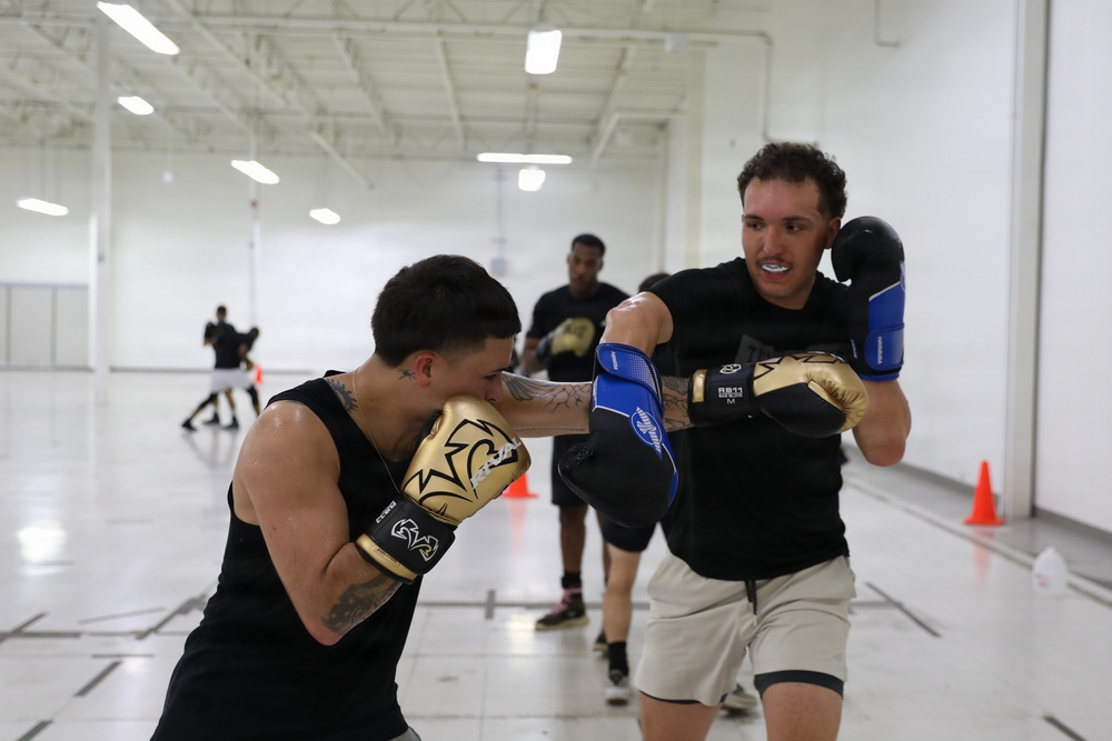 Two individuals are sparring in what appears to be an indoor gymnasium or training facility. The foreground boxer wearing gold gloves is throwing a punch while the opponent in blue and black gloves is in a defensive stance. Additional participants and orange traffic cones are visible in the background, suggesting an organized boxing training session.