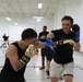 Two individuals are sparring in what appears to be an indoor gymnasium or training facility. The foreground boxer wearing gold gloves is throwing a punch while the opponent in blue and black gloves is in a defensive stance. Additional participants and orange traffic cones are visible in the background, suggesting an organized boxing training session.