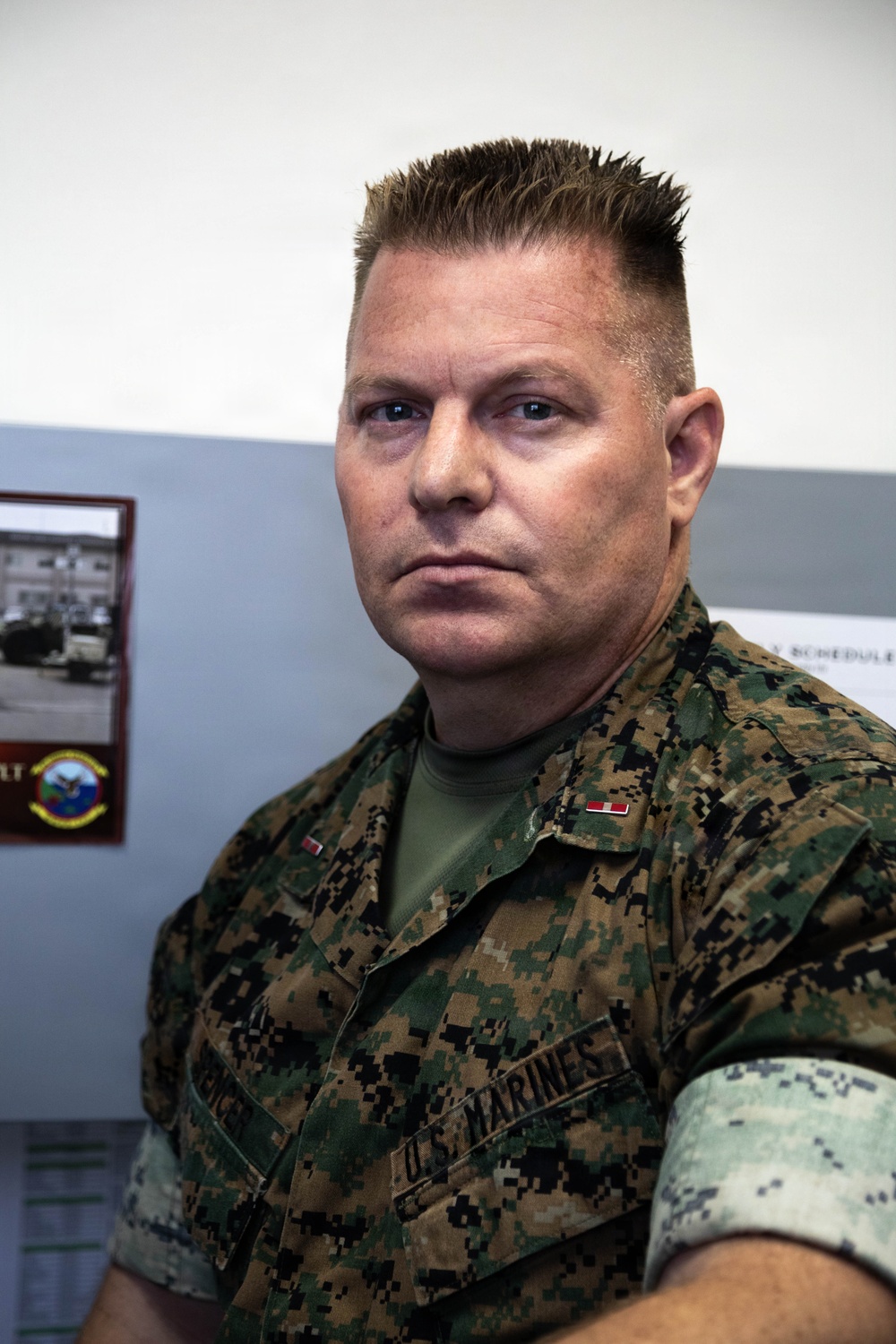 A U.S. Marine in MARPAT (Marine Pattern) digital camouflage uniform is seated in what appears to be an office or administrative space. The individual displays rank insignia on the collar and a 'U.S. MARINES' chest tape is visible. The background includes a framed unit photo/plaque and what appears to be a schedule board.