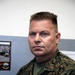 A U.S. Marine in MARPAT (Marine Pattern) digital camouflage uniform is seated in what appears to be an office or administrative space. The individual displays rank insignia on the collar and a 'U.S. MARINES' chest tape is visible. The background includes a framed unit photo/plaque and what appears to be a schedule board.