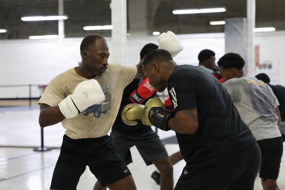 Two men are sparring in a boxing training session inside a large indoor facility. The foreground boxer wears white Venum gloves and a beige t-shirt, while his opponent wears gold-and-black Everlast gloves and a black Adidas shirt. Additional participants are visible in the background engaged in similar training activities.