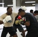 Two men are sparring in a boxing training session inside a large indoor facility. The foreground boxer wears white Venum gloves and a beige t-shirt, while his opponent wears gold-and-black Everlast gloves and a black Adidas shirt. Additional participants are visible in the background engaged in similar training activities.