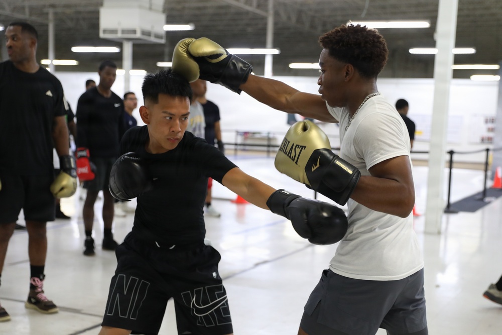 Two young men are sparring in what appears to be an indoor boxing training session. The boxer on the left wears all-black Nike gear with black gloves, while the boxer on the right wears a white shirt and gray shorts with gold-and-black Everlast gloves. Several onlookers and other participants are visible in the background of what appears to be a large gymnasium or training facility.