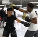 Two young men are sparring in what appears to be an indoor boxing training session. The boxer on the left wears all-black Nike gear with black gloves, while the boxer on the right wears a white shirt and gray shorts with gold-and-black Everlast gloves. Several onlookers and other participants are visible in the background of what appears to be a large gymnasium or training facility.