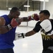 Two men are sparring in what appears to be a boxing gym or training facility with a white interior. The man on the left wears a blue sleeveless shirt and red/dark Everlast boxing gloves, while the man on the right wears a black Baltimore Ravens Football shirt and white Fighting-brand boxing gloves. Both are actively exchanging punches in a training session.