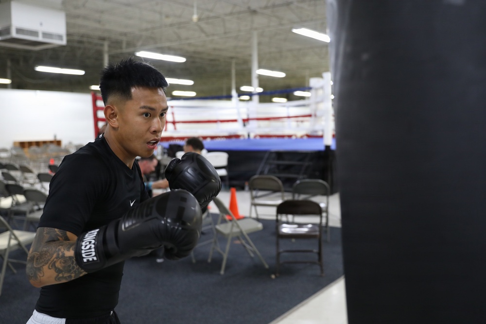A young male boxer is training at a boxing gym, striking a heavy bag with black Ringside brand boxing gloves. He is wearing a black Nike shirt and has visible tattoos on his right arm. A regulation boxing ring with red, white, and blue ropes is visible in the background along with folding chairs and other gym equipment.