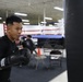 A young male boxer is training at a boxing gym, striking a heavy bag with black Ringside brand boxing gloves. He is wearing a black Nike shirt and has visible tattoos on his right arm. A regulation boxing ring with red, white, and blue ropes is visible in the background along with folding chairs and other gym equipment.
