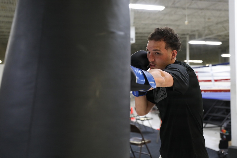 A male boxer is training on a heavy bag in what appears to be a professional boxing gym. He is wearing blue and black boxing gloves and a black athletic shirt, captured mid-punch with focused intensity. A boxing ring is visible in the background of the well-lit indoor facility.