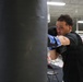 A male boxer is training on a heavy bag in what appears to be a professional boxing gym. He is wearing blue and black boxing gloves and a black athletic shirt, captured mid-punch with focused intensity. A boxing ring is visible in the background of the well-lit indoor facility.