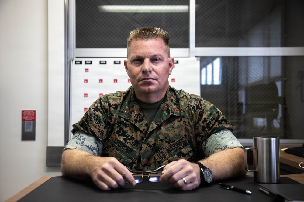 A U.S. Marines service member in MARPAT woodland camouflage uniform is seated at a desk in what appears to be an office or administrative setting. He is holding a pair of glasses and looking directly at the camera with a composed expression. A weekly calendar board and security mesh windows are visible in the background.