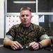 A U.S. Marines service member in MARPAT woodland camouflage uniform is seated at a desk in what appears to be an office or administrative setting. He is holding a pair of glasses and looking directly at the camera with a composed expression. A weekly calendar board and security mesh windows are visible in the background.